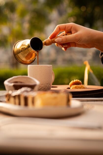 Hand pouring Turkish coffee into a cup with pastries outside, warm tones.